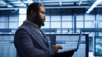 African american man walking in high tech facility, using laptop to patch network issues leading to slow data transfer rates. Data center worker fixing errors affecting supercomputers, camera A photo