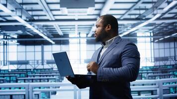 Programmer in data center typing on laptop, running error checking utilities. IT professional in server room looking for network issues affecting rigs performance using digital device, camera B photo