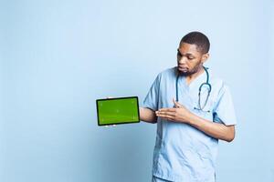 Portrait of male nurse showing medical instructions on tablet with green screen, pointing at device. Hospital employee presenting chroma key display, standing against blue background. photo