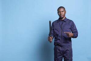 Trained security guard pointing at his baton against blue background, showing his police weapon in case of emergency. African american armed agent is on patrol and safeguarding duty. photo