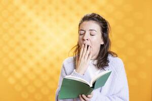 Woman yawning while reading boring lecture book needed for school exam, studio background. Student feeling fatigued while learning from academic textbook needed to pass classes photo