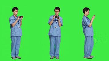 Medical assistant playing mobile games on smartphone app, taking a break from healthcare work. Nurse enjoys gaming tournament on phone, standing against greenscreen backdrop. Camera A. photo