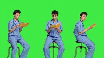 Young nurse studying for the physician exam by reading book in studio, sitting on chair against greenscreen. Specialist enjoys healthcare industry literature, preparing to become a doctor. Camera A. photo