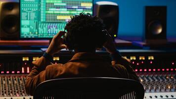 Professional tracking engineer using headphones to edit recorded tracks in control room, motorized sound control knobs. Audio expert working on mixing console and editing software. Camera A. photo