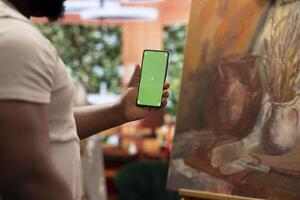 African american artist holding device with green screen mockup in art studio, working on a new painting with a certain technique. Painter using watercolor and acrylic dye for his creation. photo