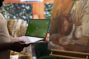 Young artist looking at isolated chroma key display in his painting atelier, using device next to canvas with a colorful masterpiece. Painter creating a rustic artwork for the gallery exhibition. photo