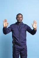 Security agent raising his hands up as a stop symbol in studio, restricting the access with a forbidden sign against blue background. Serious bodyguard in uniform does a limitation gesture. photo