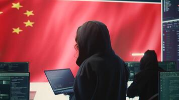 Chinese hacker working in governmental hacking room with Chinese flag on a big screen. Having the mission on inflicting fear and misinformation among the population. Dictatorship propaganda. Camera B. photo