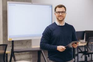 Businessman holding tablet and smiling in modern office with blank interactive whiteboard ready for presentation photo