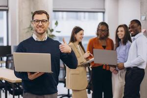 Professional working on laptop, giving thumbs up with multicultural team collaborating behind in contemporary workplace setting photo