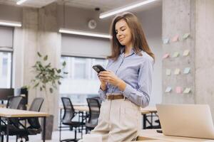 Businesswoman using a smartphone while seated at a modern office desk, working on a laptop in a bright, well designed workspace photo