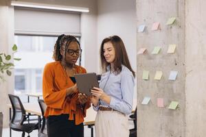 Two businesswomen are using a tablet and discussing work next to a concrete pillar with colorful sticky notes in a modern office photo