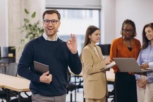 Smiling businessman showing ok sign holding tablet with diverse colleagues working on laptop in modern office, teamwork and success concept photo