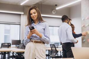 Businesswoman using mobile phone in modern office while colleagues working together on project in background photo
