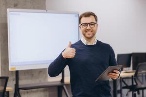 Smiling businessman showing thumbs up while holding tablet, standing in a modern office in front of blank interactive whiteboard photo