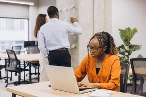Black female entrepreneur working on laptop, focusing intently while team members brainstorming nearby in modern startup workspace photo
