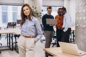 Professional businesswoman smiling confidently, standing foreground, with multicultural team collaborating on laptop project in background photo