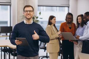 Confident businessman displaying tablet, gesturing approval with thumbs up while colleagues collaborating in contemporary workspace photo