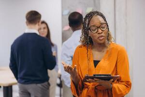 Smiling african american businesswoman holding a folder, diverse business team collaborating, analyzing data on laptops and tablets, sharing insights in contemporary workspace photo