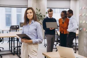 Businesswoman holding tablet smiling with multi ethnic colleagues working together using laptops in background in modern office photo