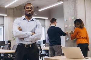 African american male with cross arms confident professional standing in an open office, while colleagues brainstorm at a wall with sticky notes, showcasing teamwork and diversity photo