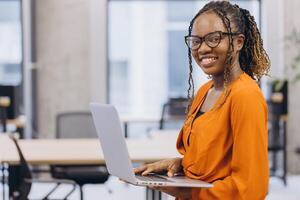 Smiling African american businesswoman with glasses working on a laptop in a contemporary office setting, showcasing diversity and modern work environments photo