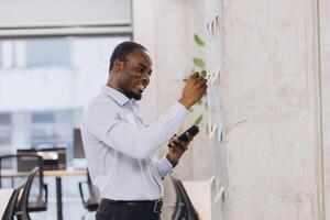 Young african american manager taking notes on colorful sticky notes attached to a wall in a modern office, using a pen and consulting his smartphone photo