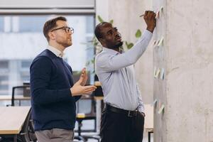 Two business professionals brainstorming and discussing ideas while writing on a wall of sticky notes in a modern office photo