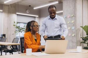 African american collaborative team analyzing data on laptop, modern workspace with greenery and brainstorming notes surrounding working colleagues photo