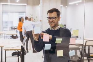 Smiling businessman using sticky notes on glass wall in a modern office, collaborating with colleagues in the background for a team project photo