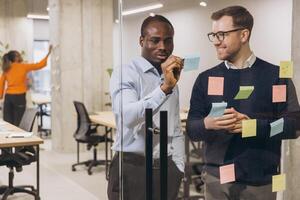 Two professional men discussing ideas with sticky notes on a glass wall in a contemporary office, promoting teamwork and innovation photo