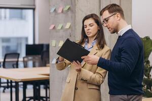 Two business professionals man and woman discussing documents in a modern office. Colorful sticky notes and a plant add vibrancy to the workspace photo