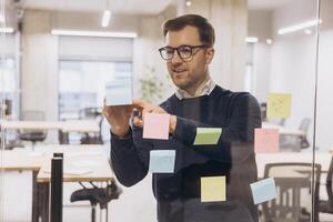 Professional man workplace team member organizing multicolored sticky notes on glass partition, collaborating during strategic planning session photo