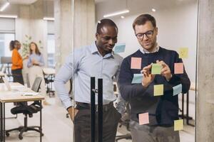 Two diverse businessmen collaborating on a project, using sticky notes on a glass wall in a modern, open plan office setting photo