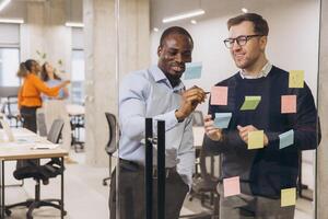 Multicultural business professionals collaborating, sharing ideas on glass wall with colorful sticky notes during strategic planning session photo