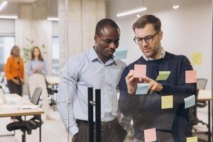 Diverse business team brainstorming, collaborating near glass wall with colorful sticky notes, showcasing innovative workplace interaction photo