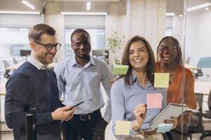 Diverse business team collaborates in a modern office, brainstorming ideas with sticky notes on glass. Smiling professionals engaged in creative discussion photo