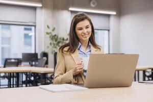 Smiling businesswoman enjoying a cup of coffee while working on a laptop in a bright, modern office space filled with natural light photo