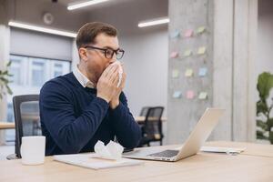 Professional businessman experiencing cold symptoms, sneezing and wiping nose while working on laptop at desk photo