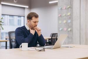 Male manager coughing while working at his desk, using a laptop in a modern office space, struggling with discomfort and fatigue photo
