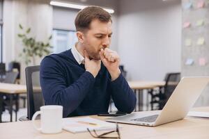 Businessman coughing and feeling unwell while working at his desk in the office, struggling with discomfort and fatigue from illness photo