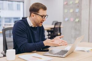 Professional businessman working remotely, gesturing during conference, using laptop at minimalist workspace with sleek design photo