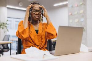 African american businesswoman experiencing stress while working on a laptop in a modern office setting, surrounded by paperwork and sticky notes photo