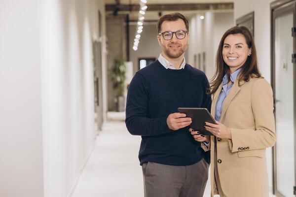 Smiling business colleagues holding a digital tablet while standing in a modern office hallway, engaging in productive discussion and collaboration