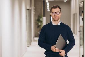 Portrait of a smiling businessman holding a laptop while walking in a bright office corridor photo
