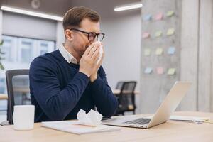 Professional working with laptop, experiencing illness, wiping nose with tissue amid office documents photo