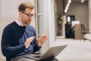 Businessman gesturing during a call on a laptop, seated in an office corridor, showcasing professional communication and focus photo