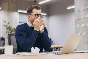 Professional working in office, blowing nose into tissue while seated at desk, battling seasonal illness photo
