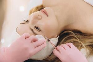 Beautician wearing pink gloves injecting botox with a syringe in forehead of young woman lying on a bed in a beauty clinic photo