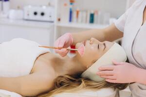 Beautician applying a pink mask on the face of a young woman lying on a massage table in a beauty salon photo